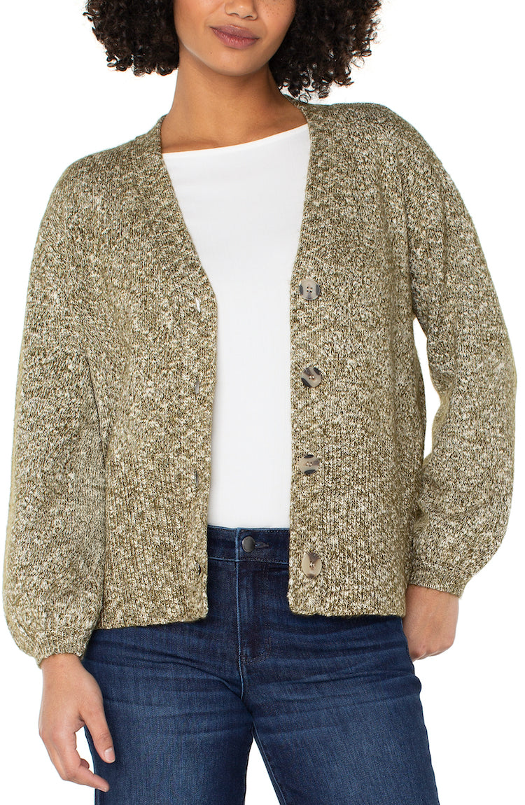 Woman wearing a textured cardigan over a white shirt and blue jeans on a plain background